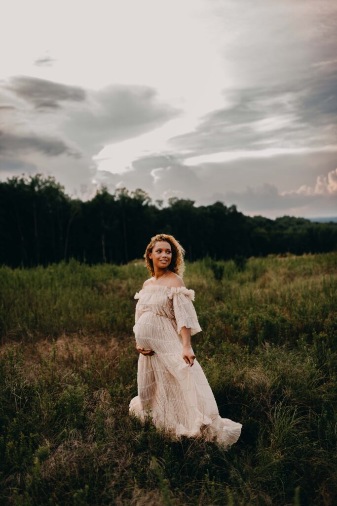 A black pregnant woman wearing a reclamation gown standing in a field with clouds.